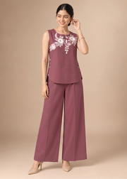 A woman modeling a sleeveless mauve jumpsuit with white floral embroidery on the chest, paired with matching wide-leg trousers and beige heels, against a solid beige studio background.