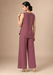 A woman modeling a sleeveless mauve jumpsuit with wide legs, shown from the back against a solid beige studio background.