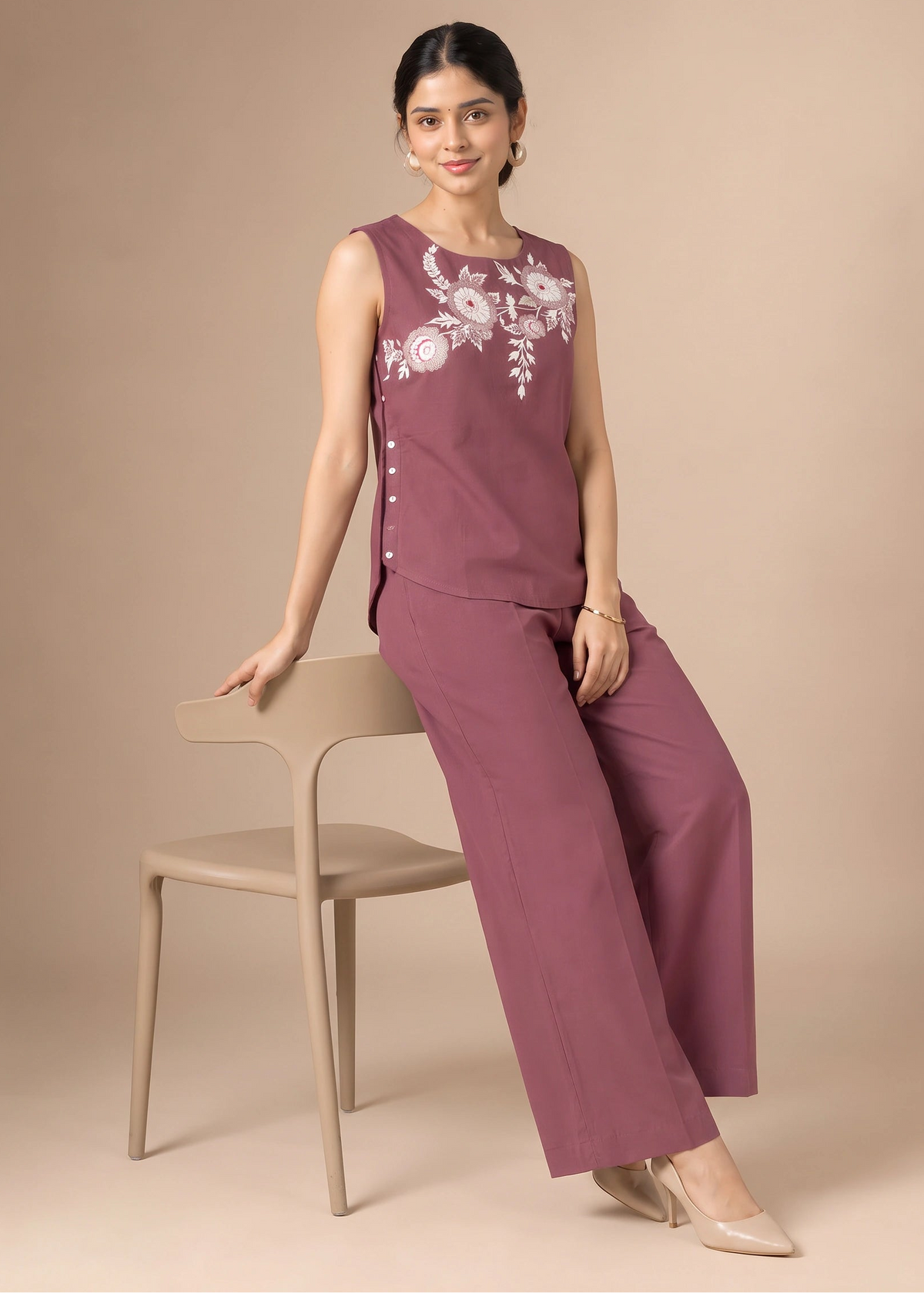 A woman models a sleeveless mauve-purple jumpsuit with white floral embroidery on the chest, paired with matching wide-leg trousers, seated on a beige chair against a solid beige background.
