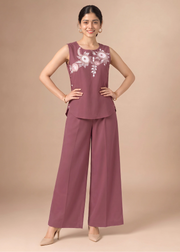 A woman modeling a sleeveless mauve jumpsuit with white floral embroidery on the chest, paired with matching wide-leg trousers, against a solid beige studio background.