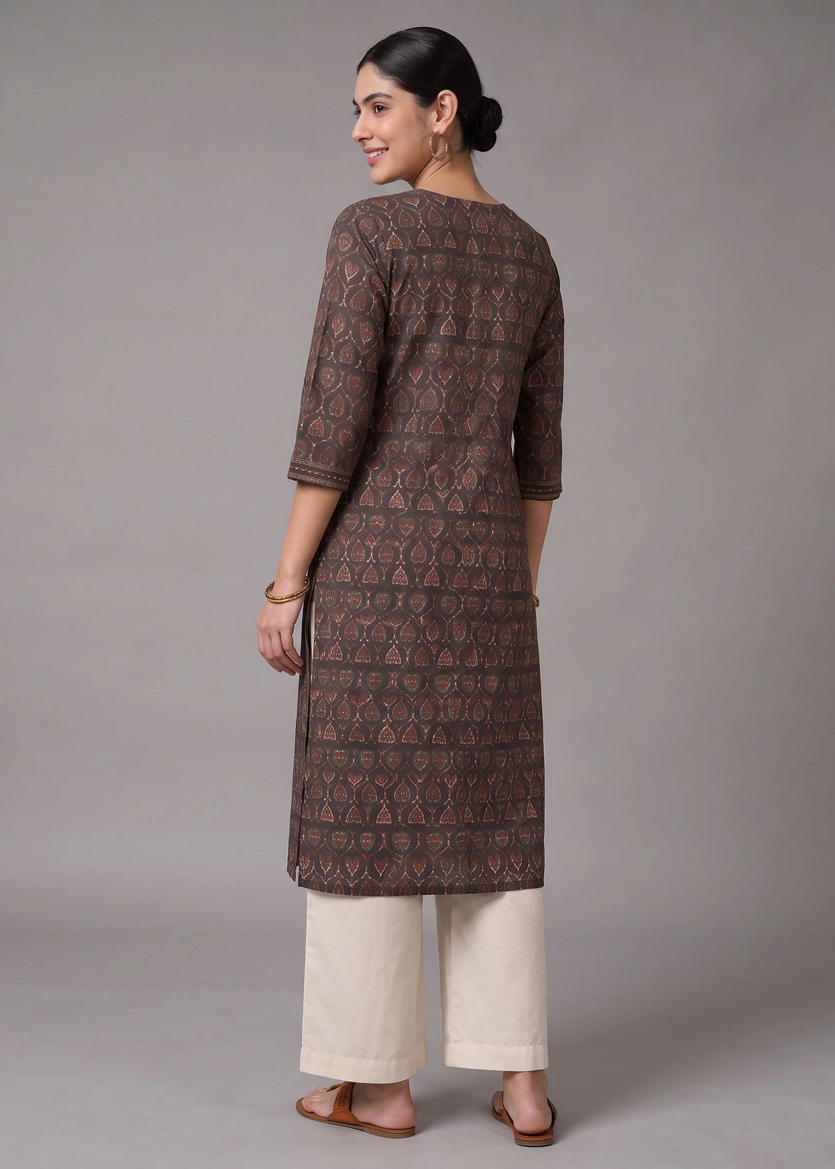 A woman stands with her back to the camera wearing a brown patterned kurta with three-quarter sleeves and cream-colored cropped pants, set against a plain gray studio background.