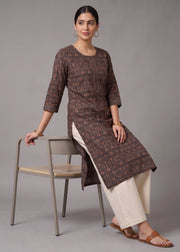 A woman models a brown printed kurta with three-quarter sleeves and a side slit, paired with off-white wide-leg pants and brown sandals, while sitting on a minimalist gray chair against a solid gray studio background.