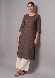 A woman modeling a brown patterned kurta with three-quarter sleeves, paired with off-white wide-leg pants and brown sandals, standing against a neutral gray studio background.
