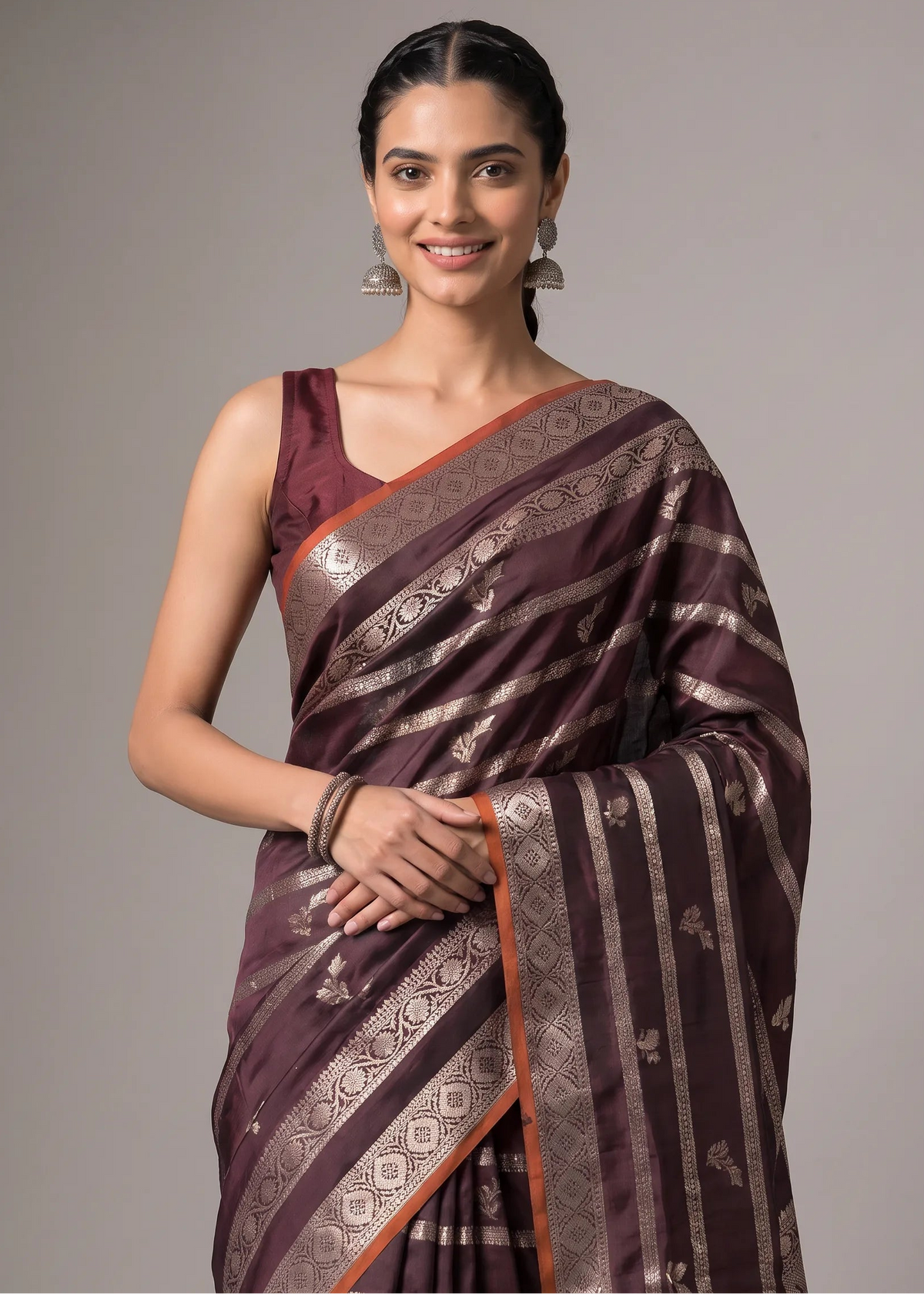 A woman modeling a dark plum purple Banarasi silk saree with metallic gold zari stripes and border patterns, paired with a matching sleeveless blouse, set against a solid gray studio background.