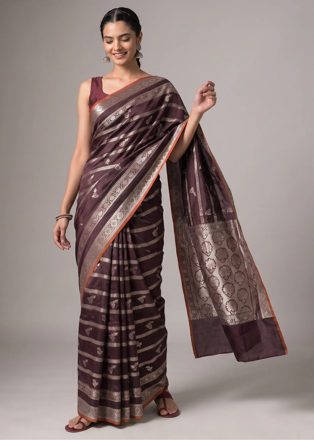 A woman models a dark brown and gold patterned silk saree with a matching sleeveless blouse against a neutral gray studio background.