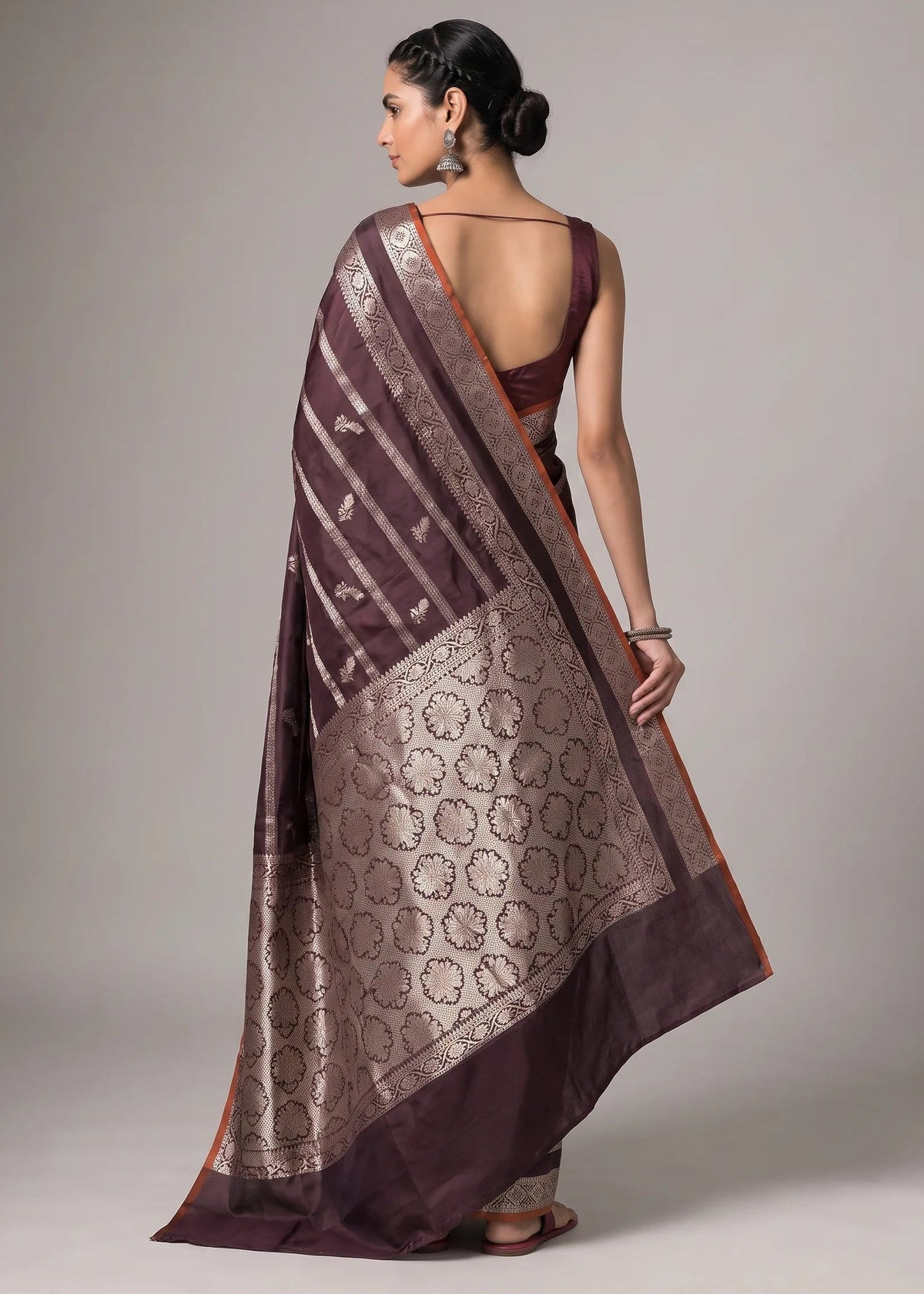 A woman modeling a dark plum purple saree with a wide metallic gold border and intricate woven patterns, shown from the back against a neutral gray studio background.