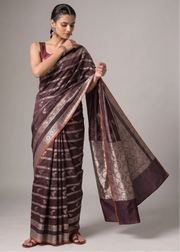 A woman modeling a dark brown and silver patterned silk saree with a matching sleeveless blouse, standing against a neutral gray studio background.