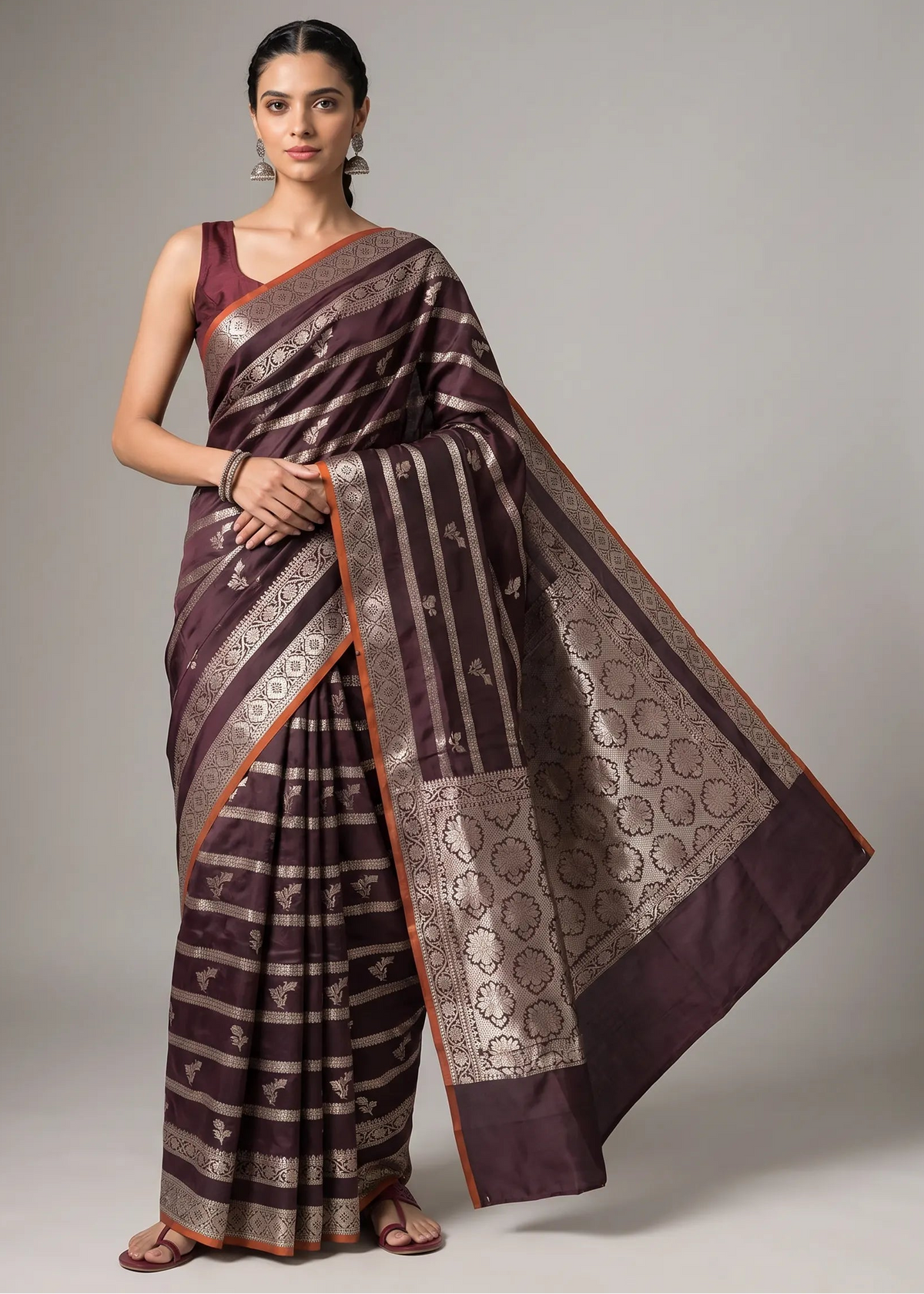 A woman modeling a dark purple and gold Banarasi silk saree with a matching purple blouse, standing against a neutral gray studio background.