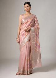 A woman modeling a coral orange and pink saree with shimmering metallic gold and silver zari patterns and a wide decorative border, photographed against a neutral gray studio background.