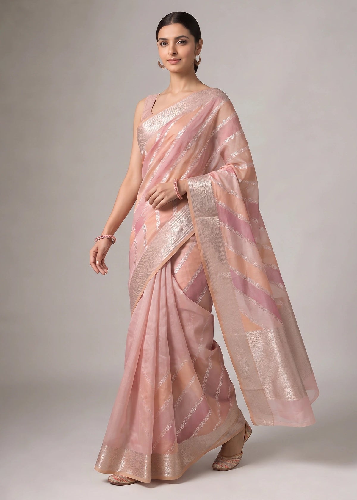 A woman modeling a coral orange and pink saree with metallic gold stripes, posed against a neutral gray studio background.