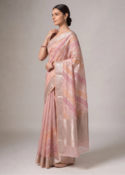 A woman modeling a coral orange and pink saree with metallic silver stripes and a wide silver border, posed against a neutral gray studio background.
