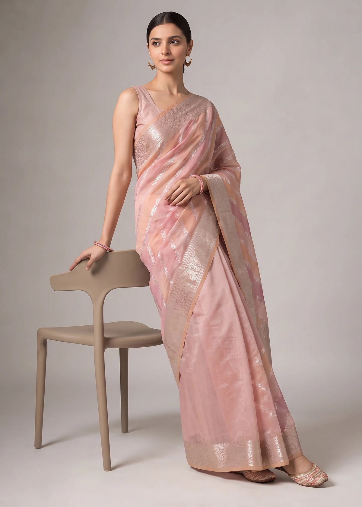 A woman models a coral orange and pink saree with a shimmering metallic border, standing next to a minimalist beige chair against a neutral gray studio background.