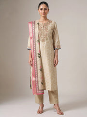 A woman modeling a traditional beige salwar kameez suit with floral embroidery, matching trousers, and a pink patterned dupatta, set against a plain beige studio background.
