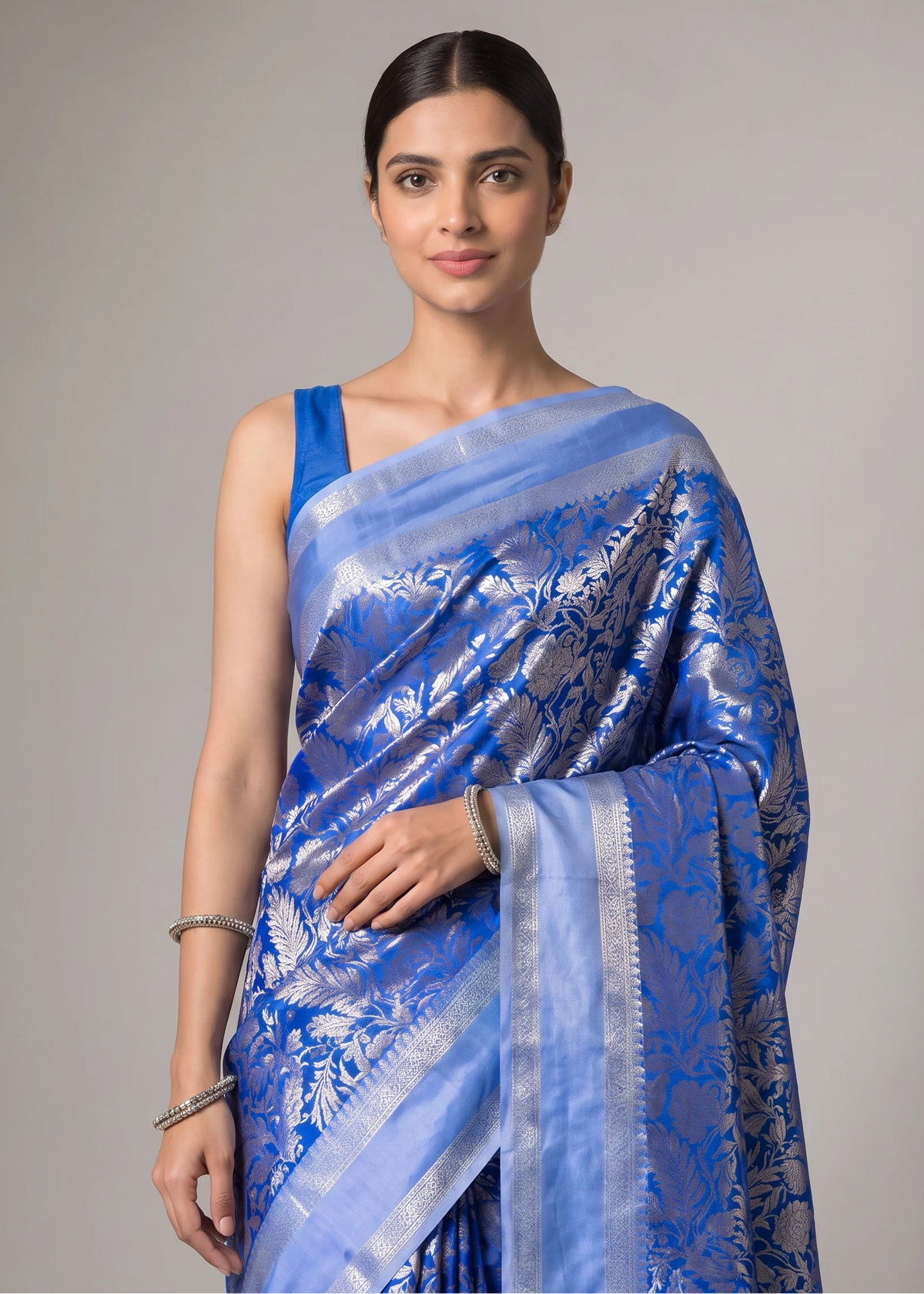 A woman modeling a blue and silver floral Banarasi silk saree with a matching blue blouse, set against a solid gray studio background.