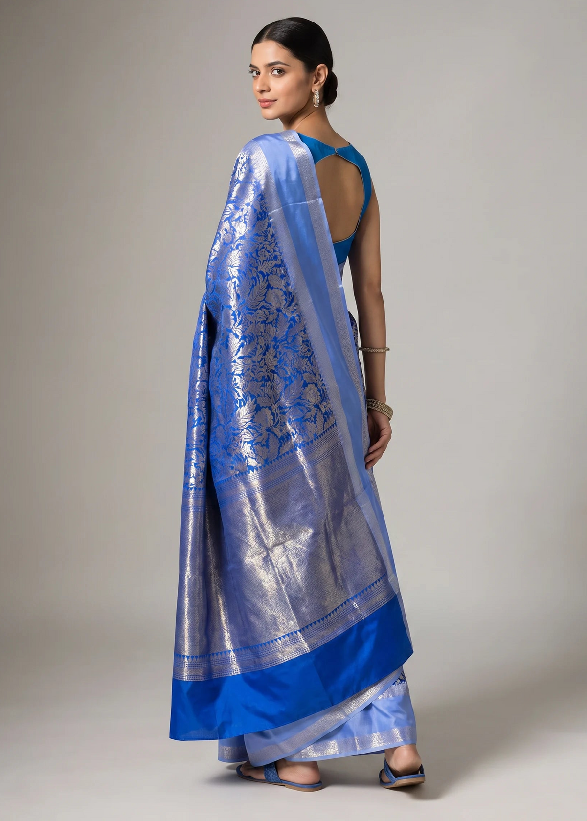 A woman modeling a royal blue and silver Banarasi silk saree with intricate floral zari embroidery, shown from a three-quarter back view against a neutral studio background.