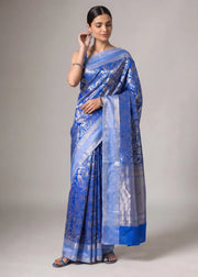 A woman modeling a royal blue and silver patterned silk saree against a neutral gray studio background.