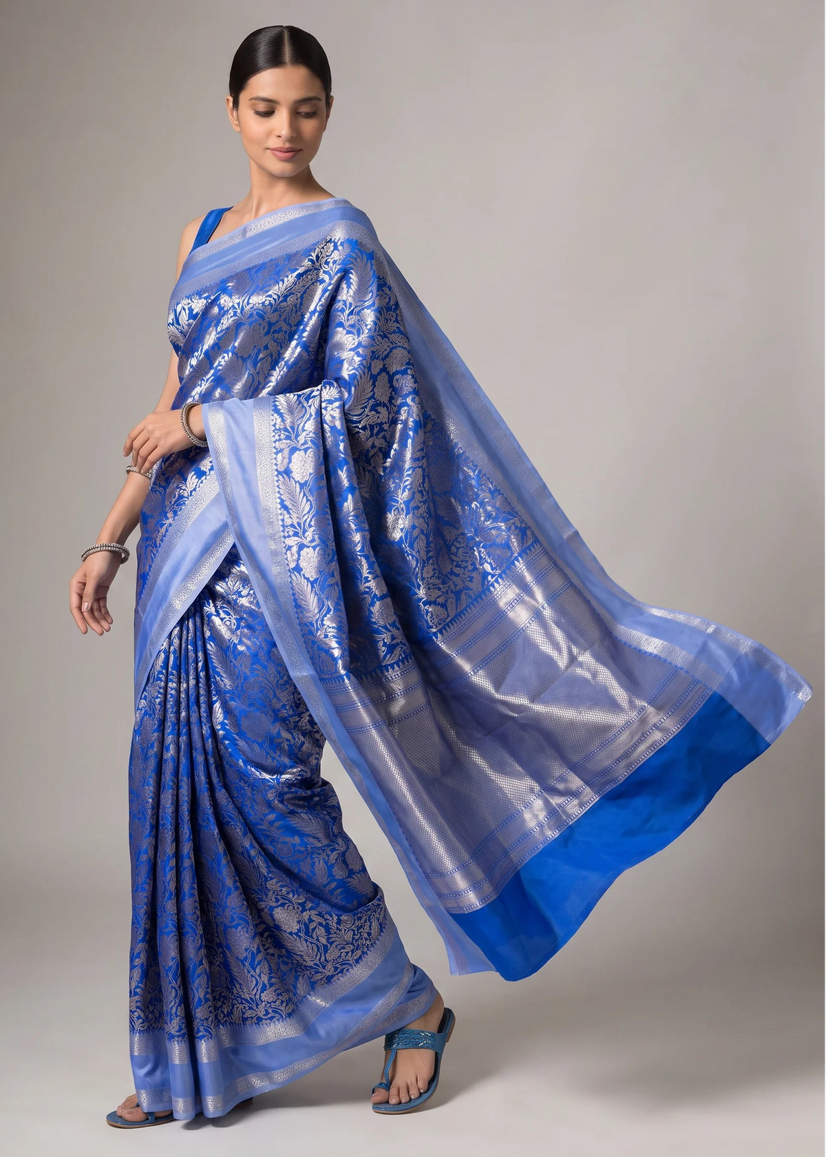 A woman modeling a blue and silver patterned silk saree against a neutral gray studio background.