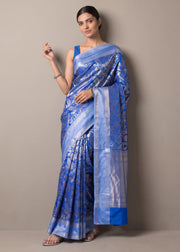 A woman modeling a blue and silver patterned silk saree with a matching blue blouse, standing against a plain beige studio background.