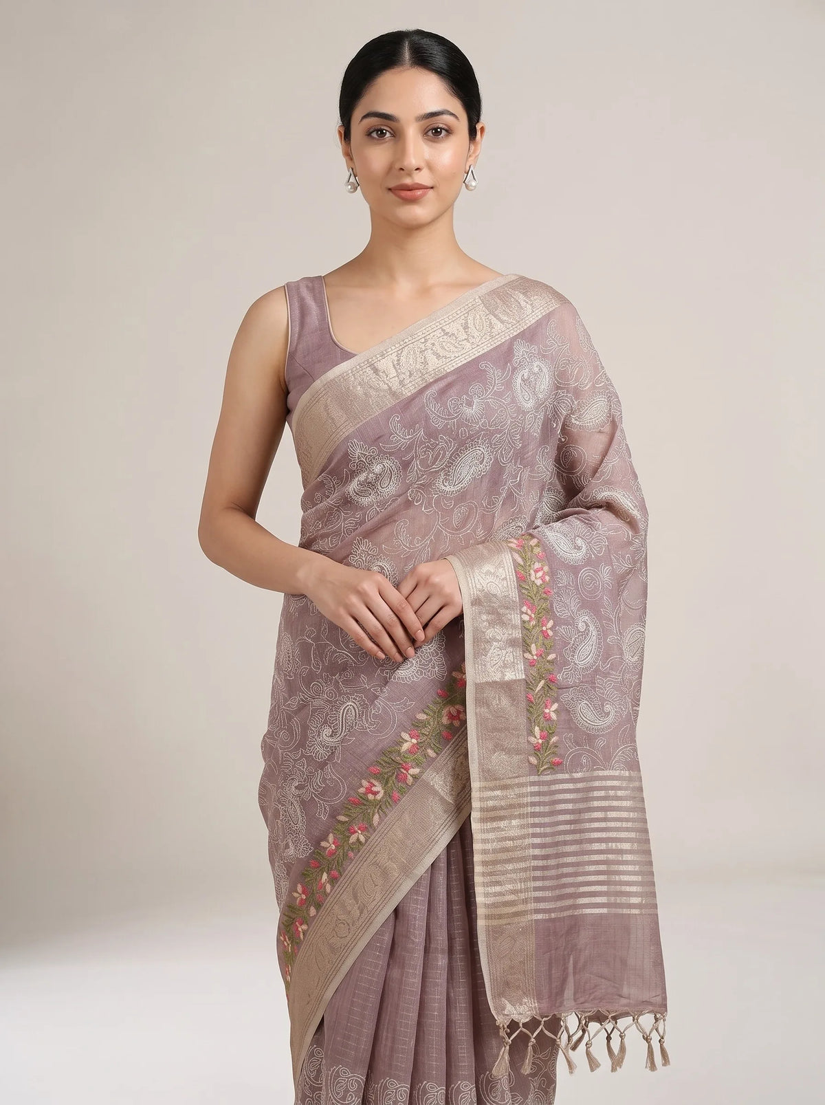 A woman modeling a draped lavender-gray silk saree with beige borders, paisley motifs, and pink floral embroidery, set against a plain light beige studio background.