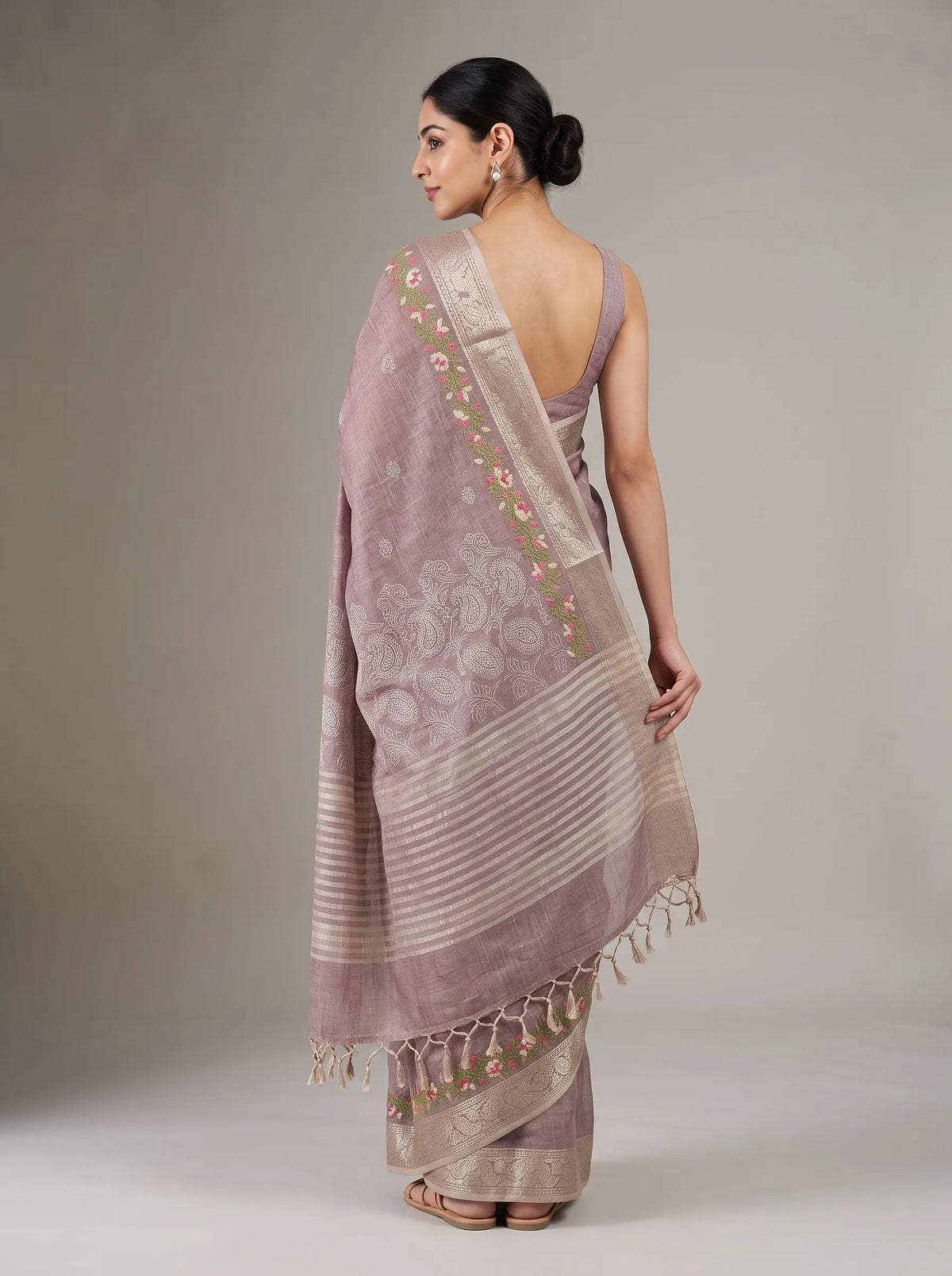 A woman modeling a dusty lavender-pink saree with beige stripes, floral motifs, and a matching blouse against a neutral studio background.