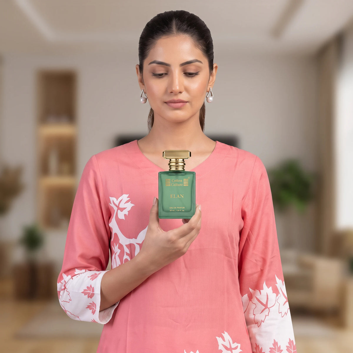 A woman in a pink kurta holds a green rectangular perfume bottle with a gold cap. The background is a softly blurred indoor living room setting.