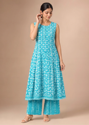 A woman modeling a sleeveless turquoise blue and white printed kurta with matching wide-leg pants, standing against a solid beige studio background.