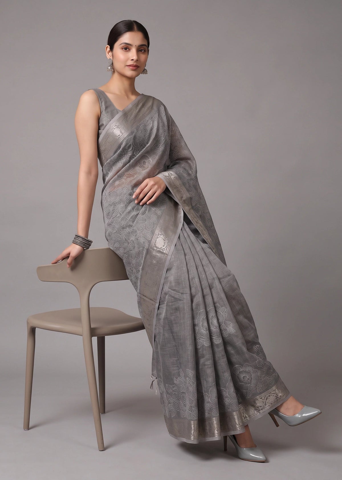 A woman models a metallic silver-gray saree with subtle floral motifs and a matching border, seated on a minimalist beige chair against a solid gray studio background.