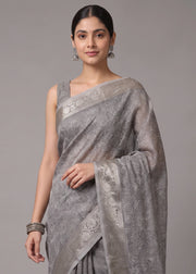 A woman modeling a light gray semi-sheer saree with intricate silver floral embroidery and a matching sleeveless blouse, set against a solid gray studio background.