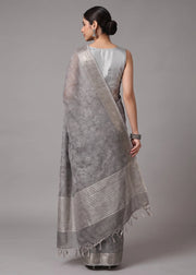 A woman modeling a light gray saree with a silver floral patterned body and a striped border, shown from the back against a solid gray studio background.