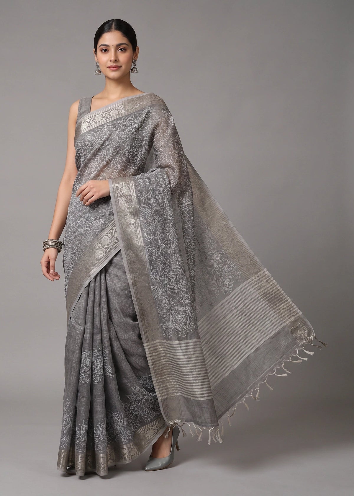 A woman models a light gray saree with silver-toned floral patterns and a matching dupatta, standing against a solid gray studio background.