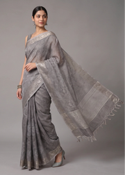 A woman modeling a light gray saree with subtle floral patterns and a matching dupatta, posed against a solid gray studio background.