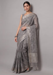 A woman modeling a gray patterned saree with a silver-toned border and pallu, standing against a plain gray studio background.
