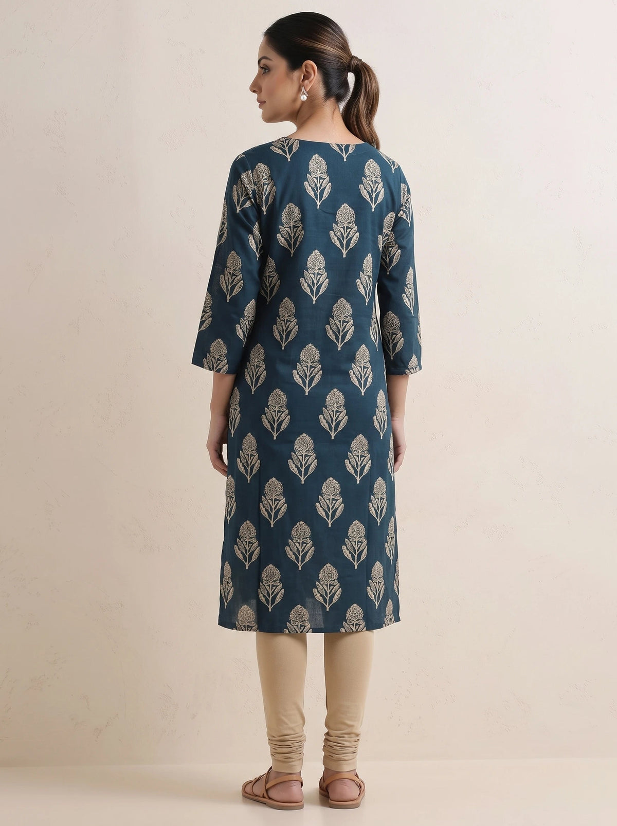 A woman shown from the back wearing a teal blue kurta with a beige leaf pattern, paired with beige leggings and brown sandals, standing against a plain beige background.