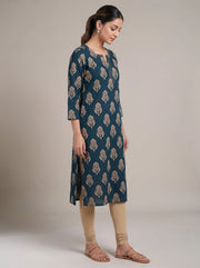 A woman modeling a dark teal blue-green kurta with a beige leaf block print, paired with beige leggings and brown sandals, standing against a neutral beige studio background.