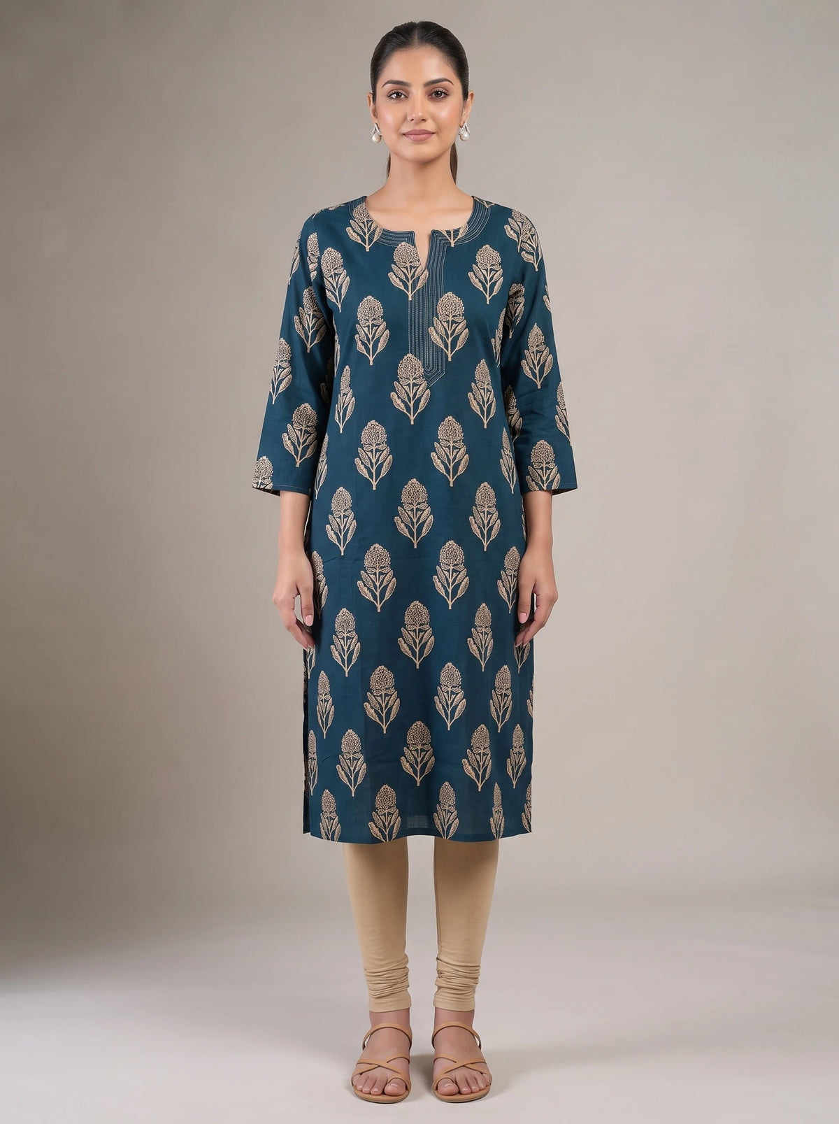A woman stands against a plain beige studio background wearing a teal-blue knee-length kurta with beige leaf-like block prints, paired with beige leggings and beige sandals.