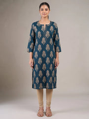 A woman stands against a plain beige studio background wearing a teal-blue knee-length kurta with beige leaf-like block prints, paired with beige leggings and beige sandals.