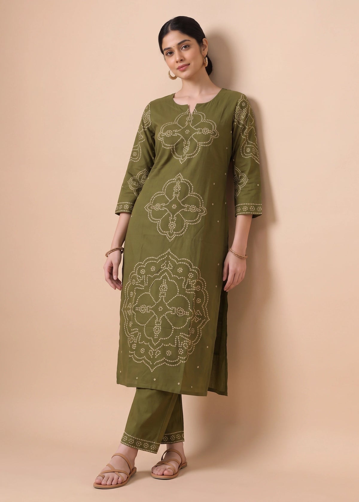 A woman modeling an olive green kurta set with beige geometric and floral mandala patterns, paired with matching pants and sandals, against a solid beige background.
