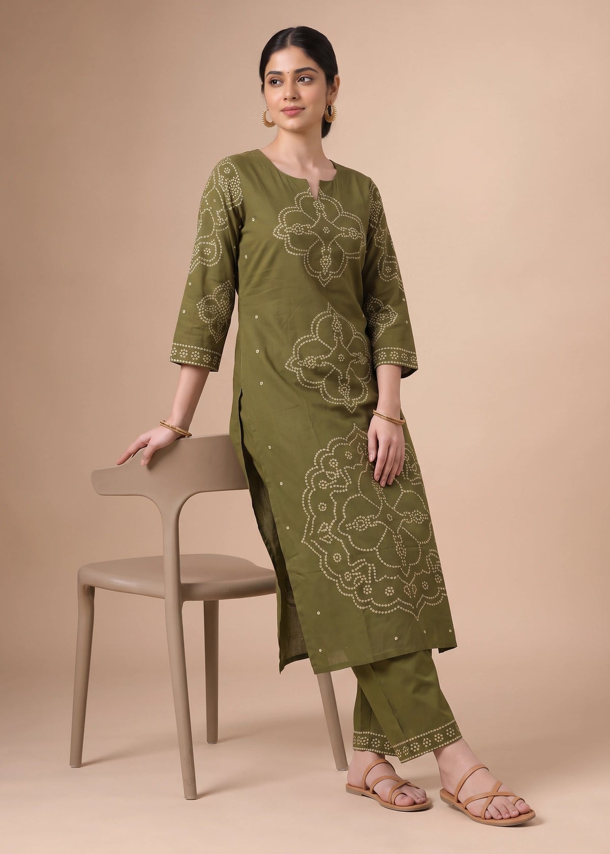 A woman models an olive green three-piece kurta set with beige floral embroidery, standing next to a beige chair against a solid beige background.