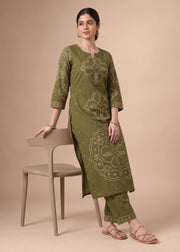 A woman models an olive green three-piece kurta set with beige floral embroidery, standing next to a beige chair against a solid beige background.