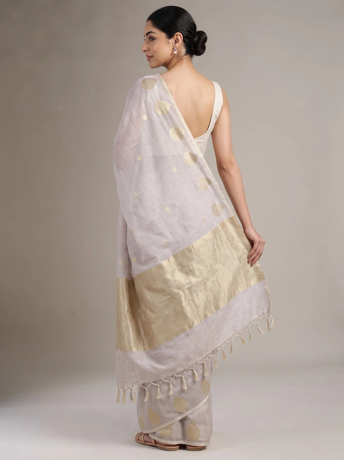 A woman modeling a lilac purple saree from the back, showing the gold foil motifs, border, and tassel fringe against a neutral studio background.