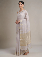 A woman modeling a lilac purple saree with gold motifs and a wide gold border, standing against a neutral beige studio background.