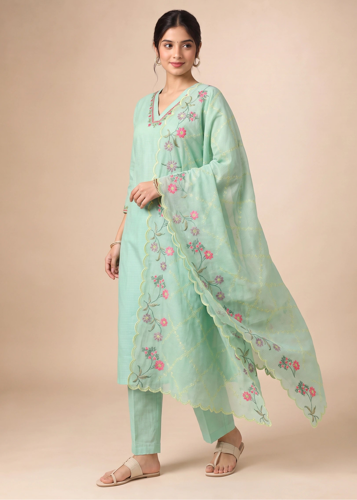 A woman modeling a mint green traditional Indian salwar kameez suit with floral embroidery, featuring a long-sleeved tunic, matching pants, and a coordinating dupatta, set against a solid beige background.