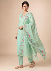 A woman modeling a mint green traditional Indian salwar kameez suit with floral embroidery, featuring a long-sleeved tunic, matching pants, and a coordinating dupatta, set against a solid beige background.