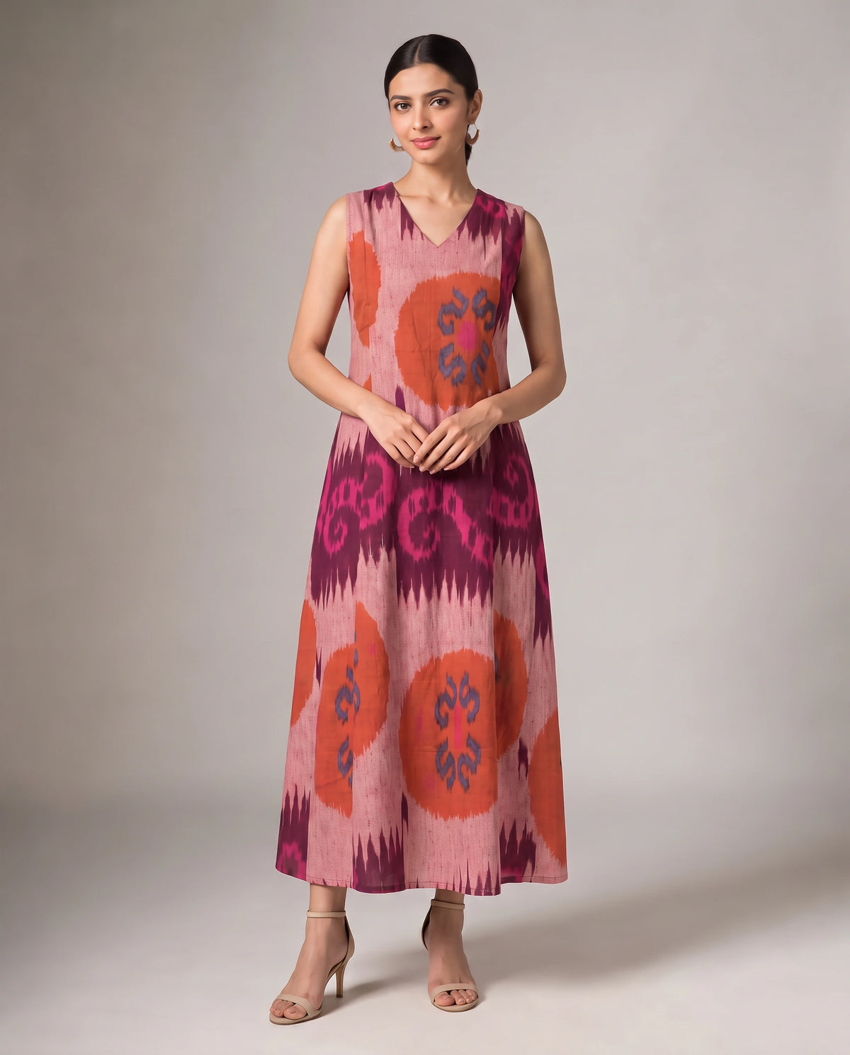 A woman modeling a sleeveless V-neck maxi dress with a pink, purple, and orange ikat pattern, standing against a neutral studio background.