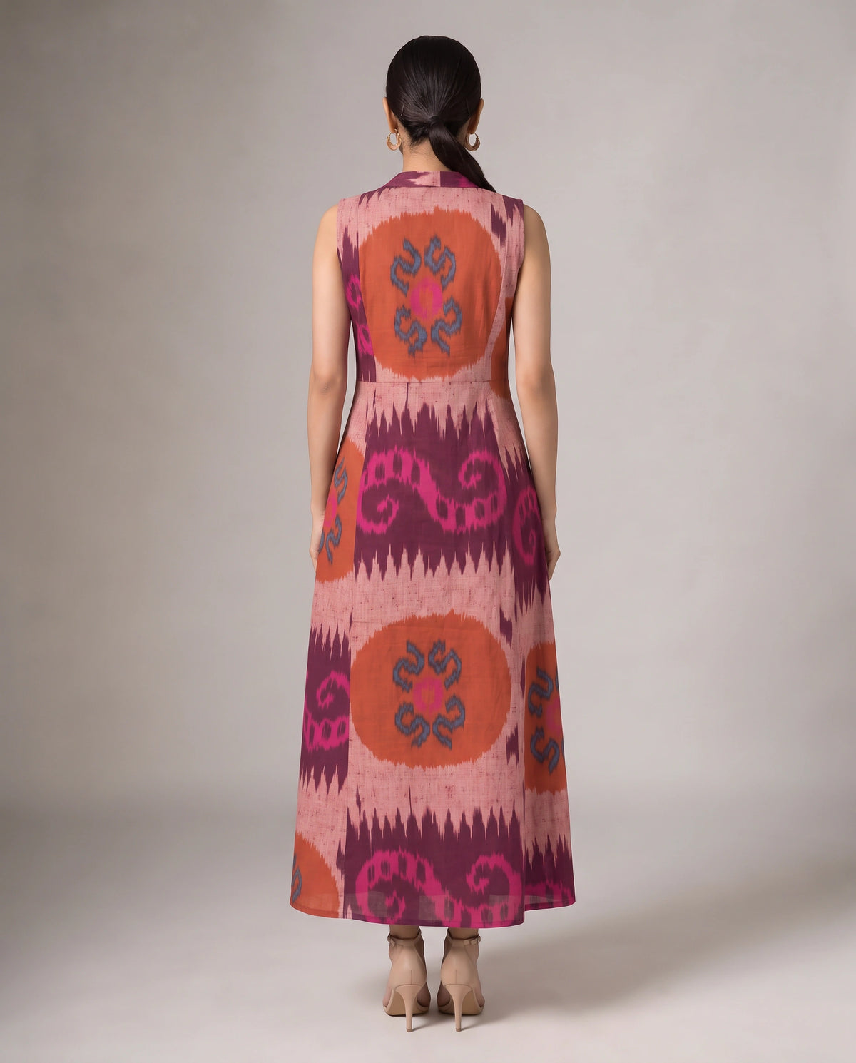 A woman viewed from the back wearing a sleeveless maxi dress with a pink, orange, and purple ikat-style pattern, standing against a neutral studio background.