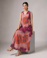 A woman models a sleeveless V-neck maxi dress with a pink, orange, and purple ikat pattern, sitting on a gray block against a neutral studio background.