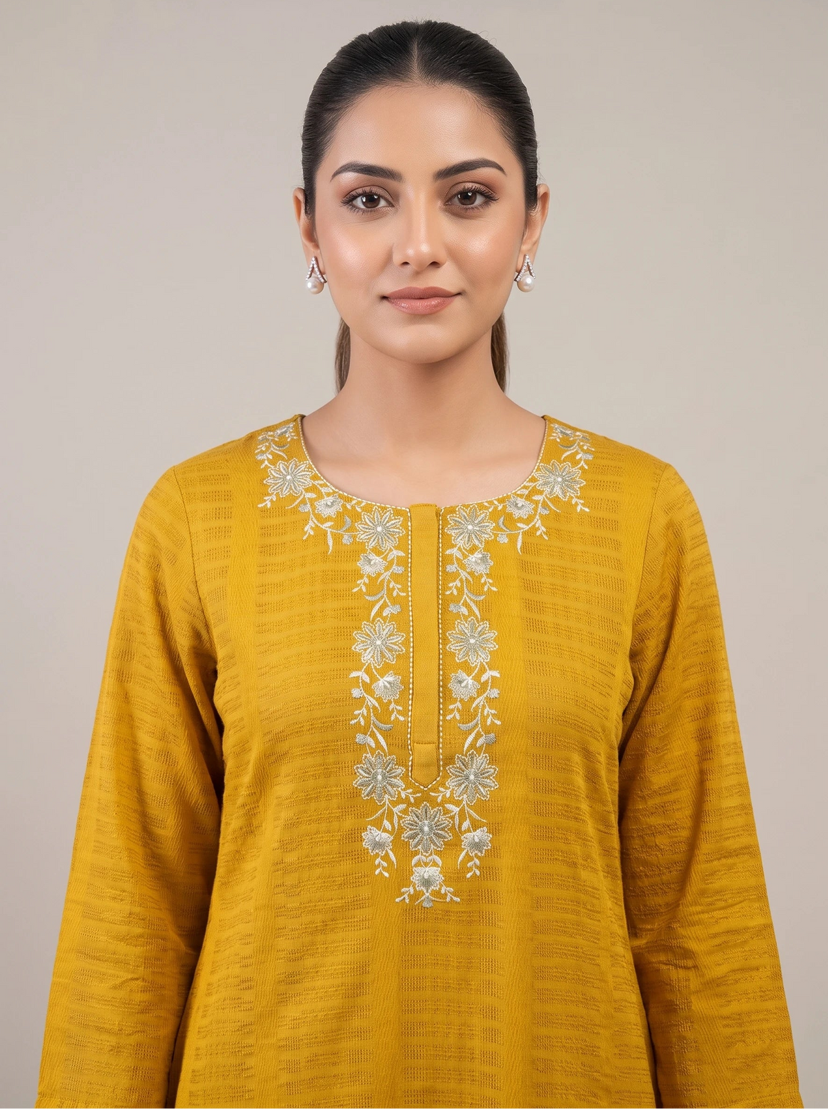 A woman wearing a mustard yellow long-sleeve kurta with intricate silver floral embroidery around the neckline, photographed against a solid light gray background.