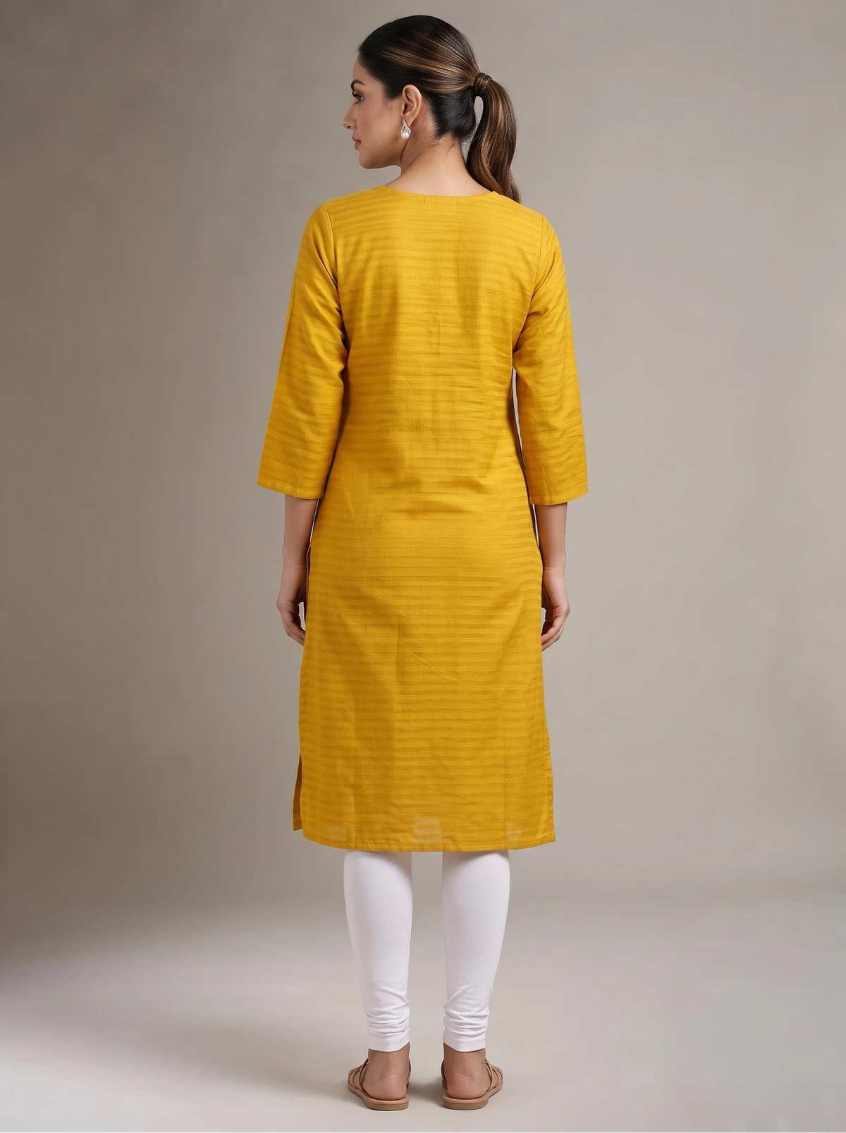 A woman viewed from the back wearing a mustard yellow textured kurta with three-quarter sleeves, paired with white leggings and brown sandals, against a neutral beige studio background.