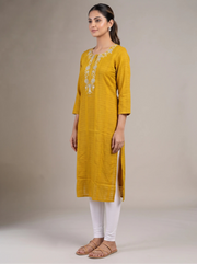 A woman modeling a mustard yellow kurta with white floral embroidery on the chest, paired with white leggings and brown sandals, set against a plain beige studio background.