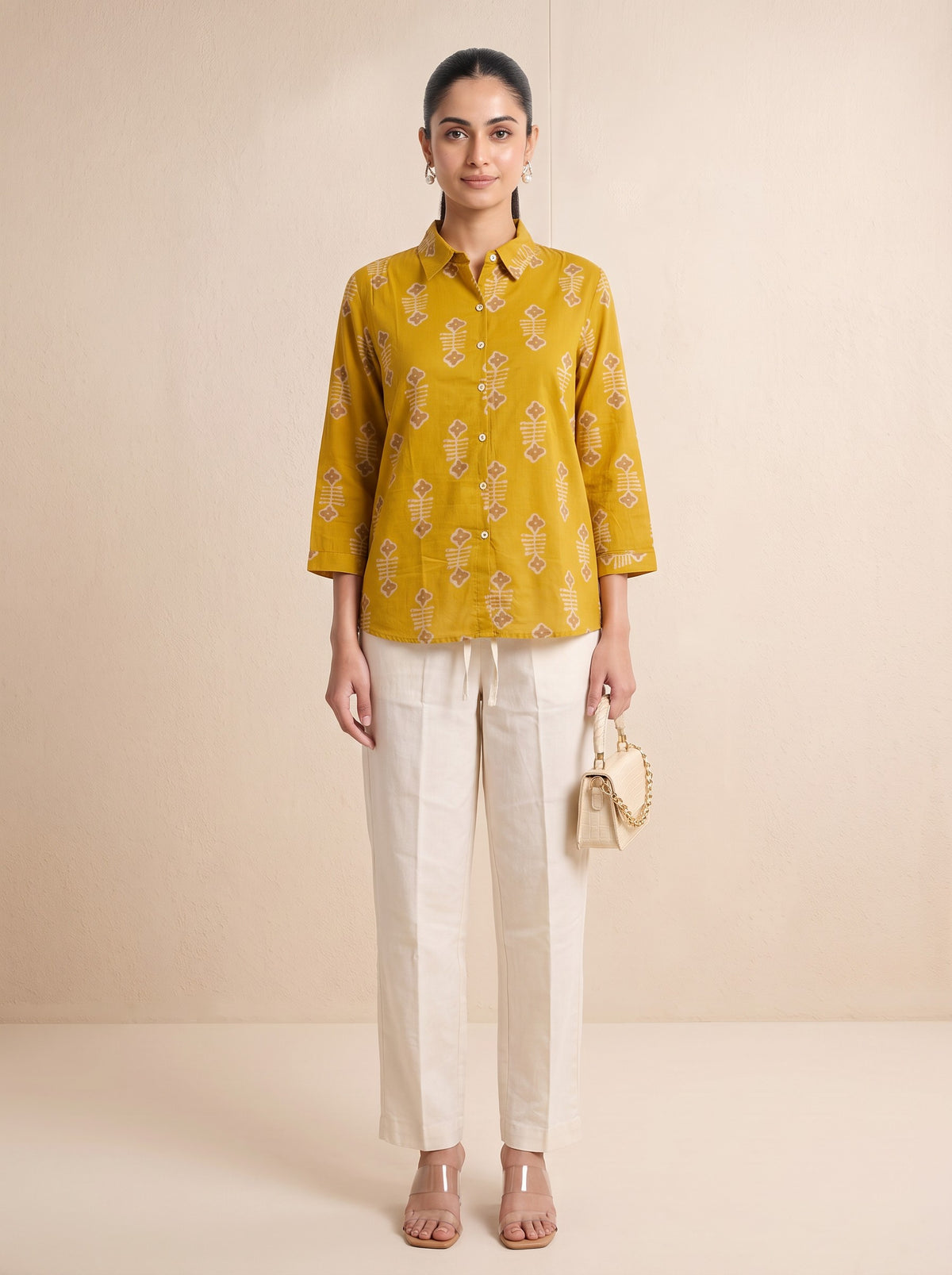 A woman stands against a plain beige studio backdrop wearing a mustard yellow printed button-up shirt with white pants, clear heeled sandals, and holding a small beige handbag.